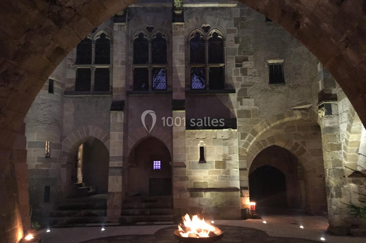 Cour intérieure en pierre éclairée par un brasero central, entourée d'arches et de fenêtres gothiques.