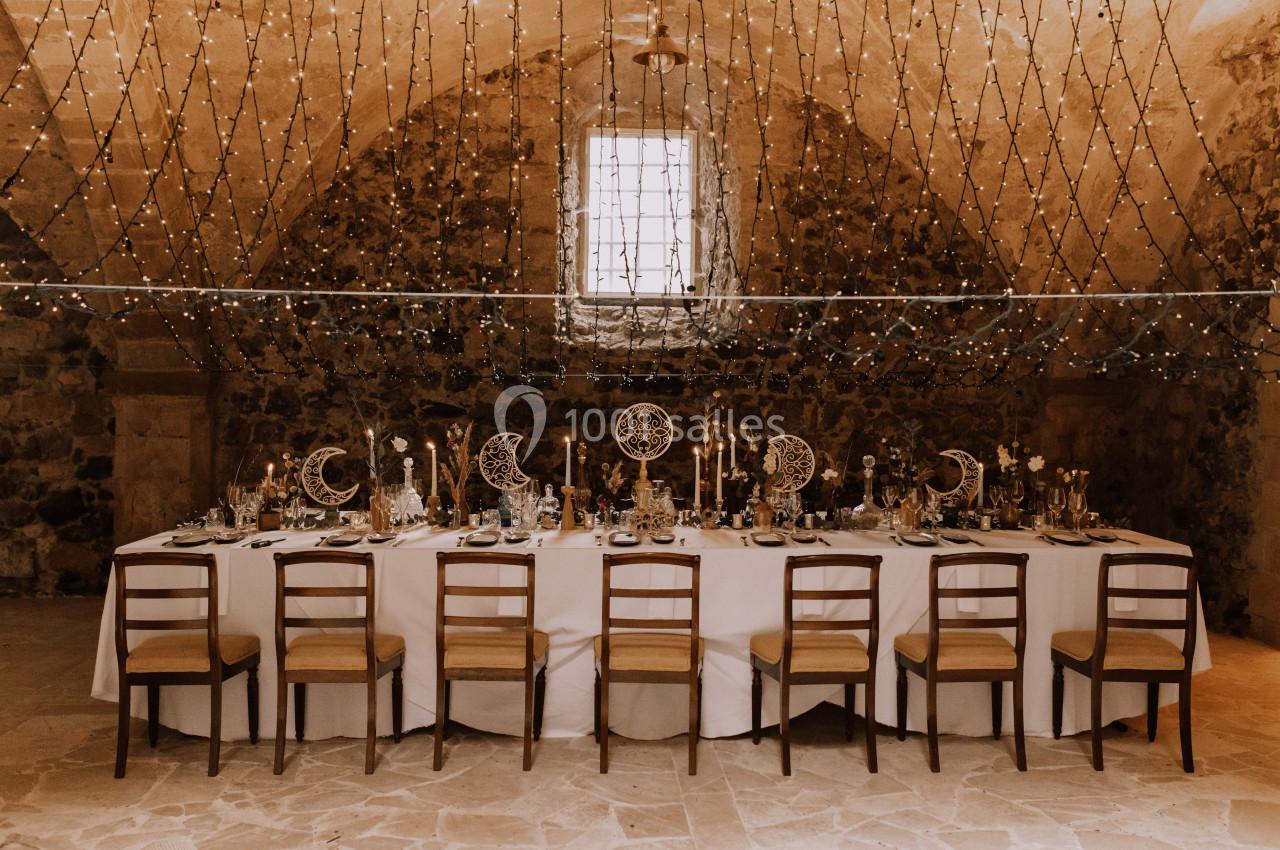 Table décorée pour un dîner dans une salle voûtée en pierre, éclairée par des guirlandes lumineuses suspendues.