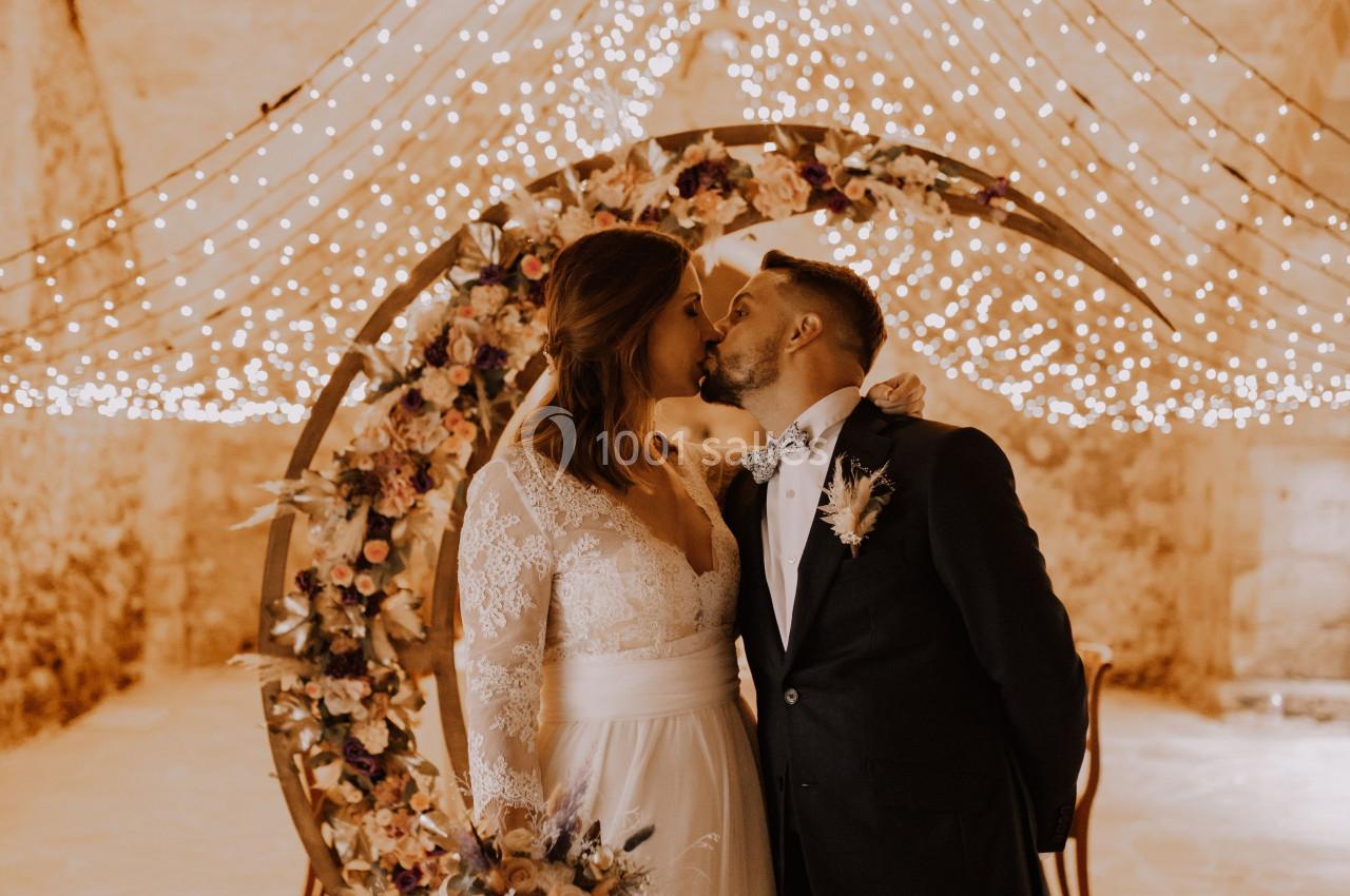 Un couple en tenue de mariage s'embrasse devant une arche décorée de fleurs et de guirlandes lumineuses.