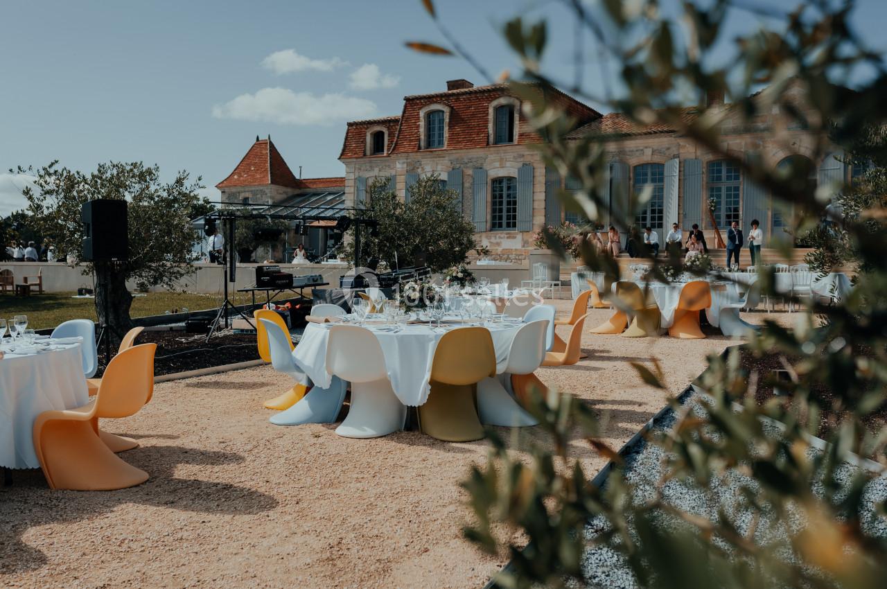 Tables dressées avec chaises colorées disposées en extérieur devant un bâtiment en pierre avec toit en tuiles rouges.