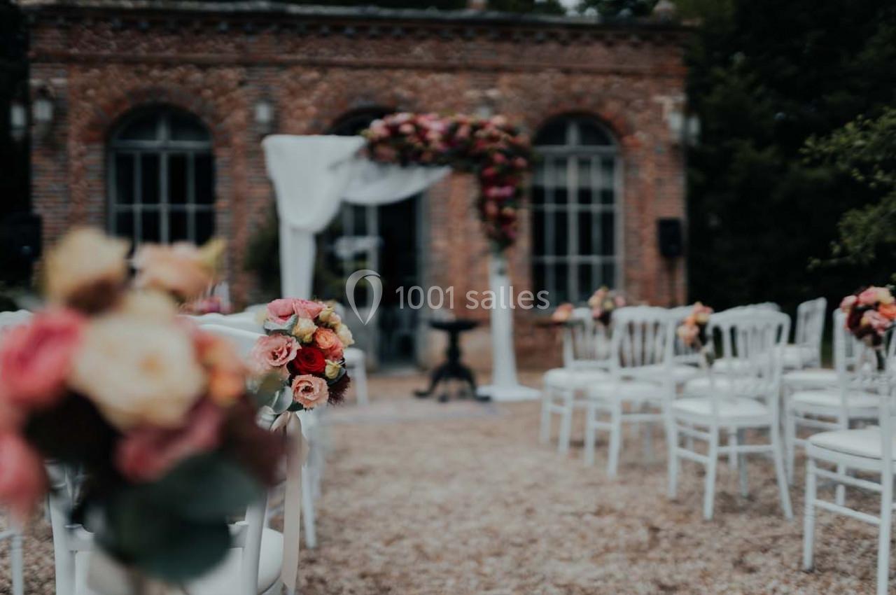 Chaises blanches alignées devant une arche fleurie dans une cour extérieure, préparée pour une cérémonie.