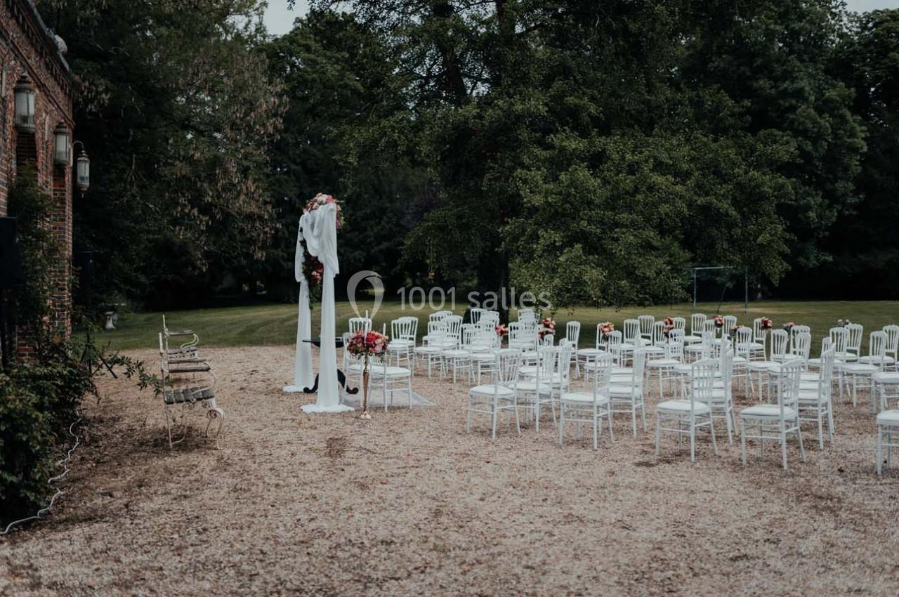 Chaises blanches disposées en extérieur devant une arche décorée de fleurs, entourées d'arbres et de verdure.