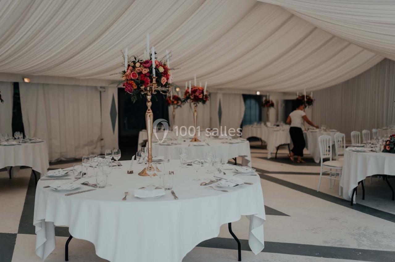 Salle de réception décorée avec des tables rondes, nappes blanches, chandeliers fleuris et une personne ajustant les détails.