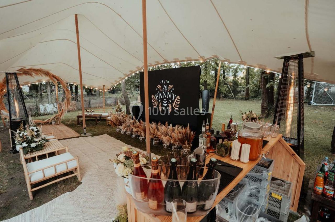 Bar extérieur décoré sous une tente avec des boissons, des fleurs et des éléments naturels dans un cadre champêtre.