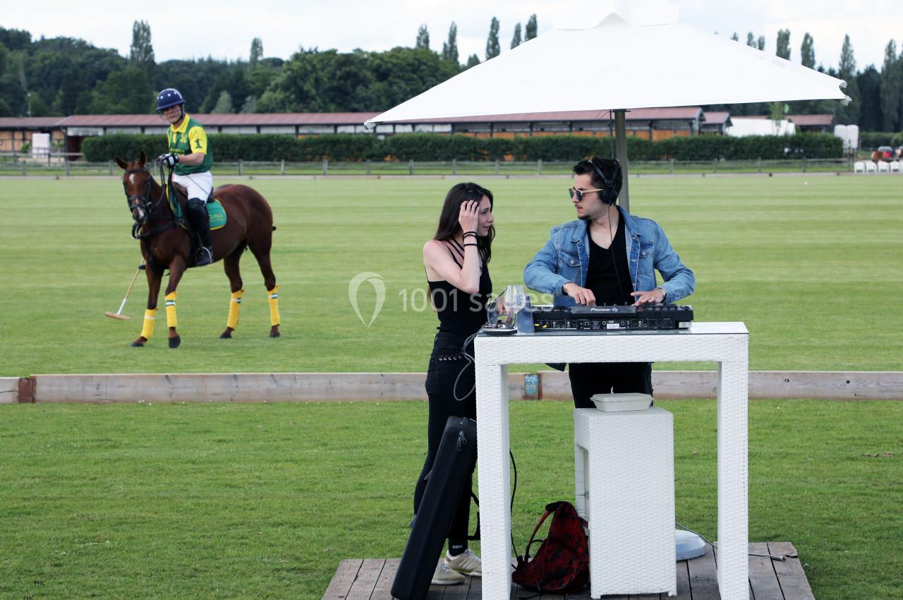 Un DJ mixe sur une pelouse près d'un terrain de polo, avec un joueur à cheval en arrière-plan.