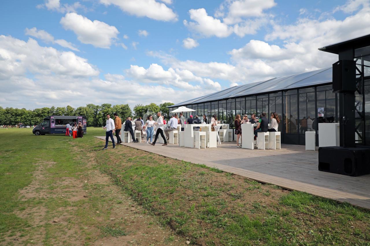 Des personnes discutent et se tiennent près de tables hautes en extérieur, à côté d'une structure vitrée et d'un food truck.