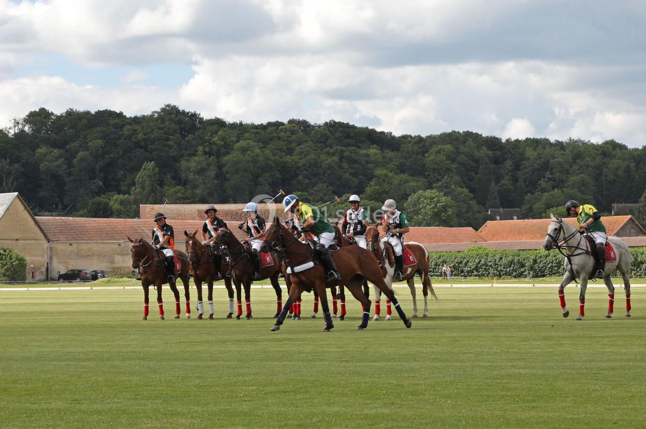Des joueurs de polo à cheval se préparent sur un terrain herbeux, avec des bâtiments et des arbres en arrière-plan.