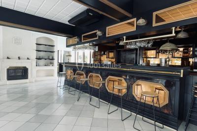 Salle lumineuse avec tables rectangulaires, chaises noires et verrerie suspendue, donnant sur de grandes fenêtres.