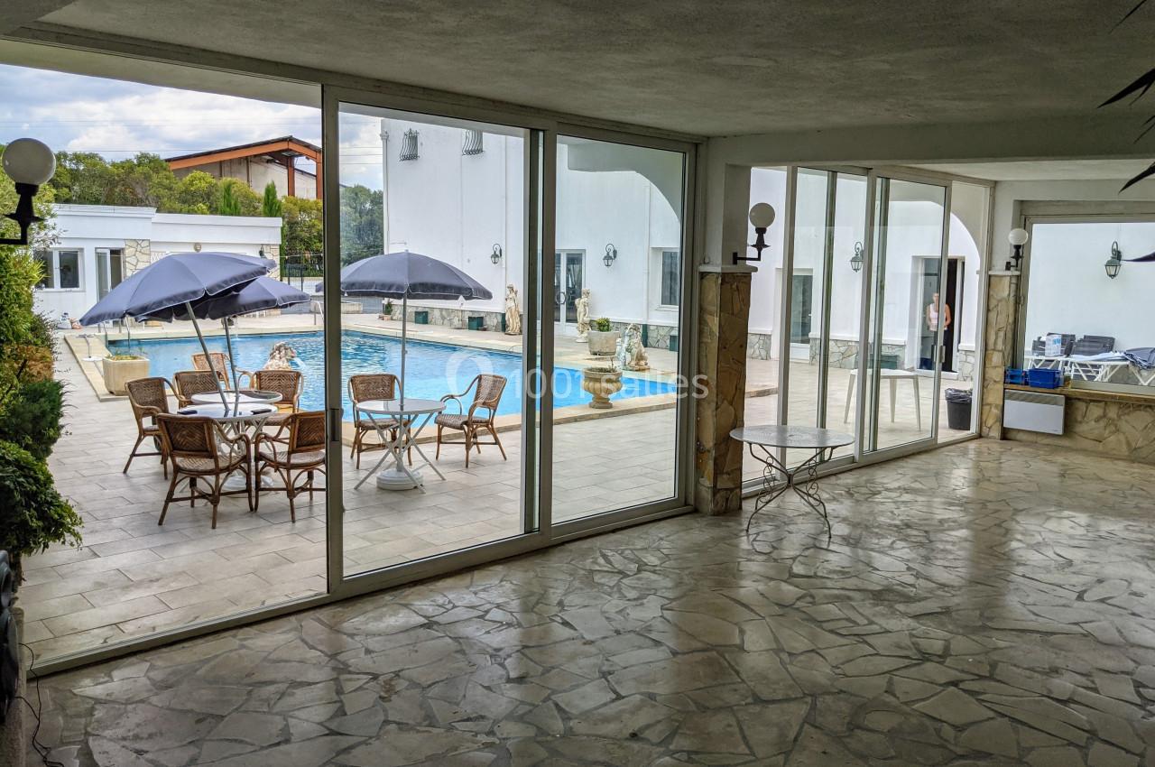 Vue depuis une pièce avec sol en pierre sur une terrasse équipée de tables, chaises, parasols et une piscine extérieure.
