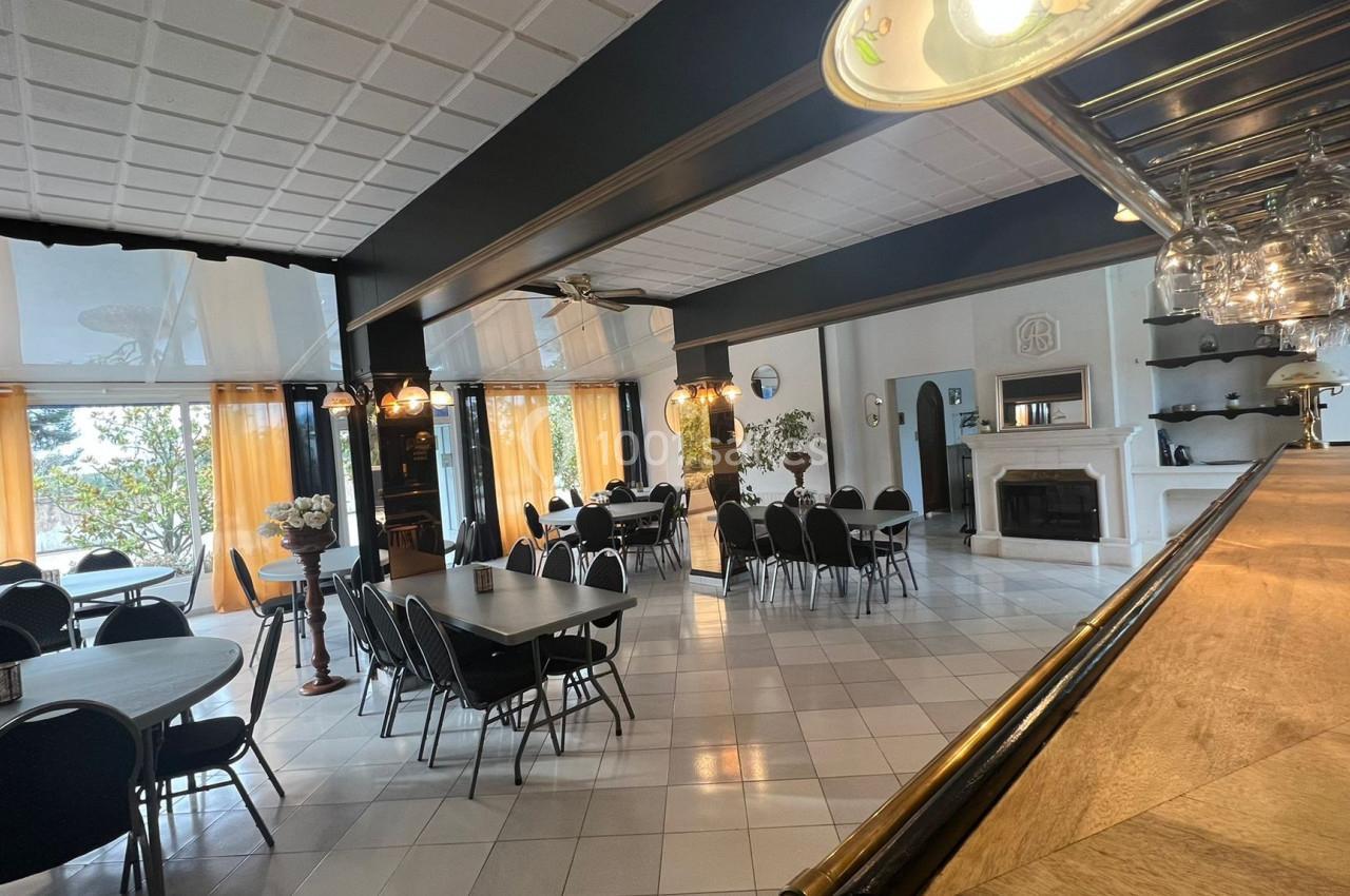 Salle lumineuse avec tables rondes et rectangulaires, chaises noires, grandes fenêtres et cheminée blanche en arrière-plan.