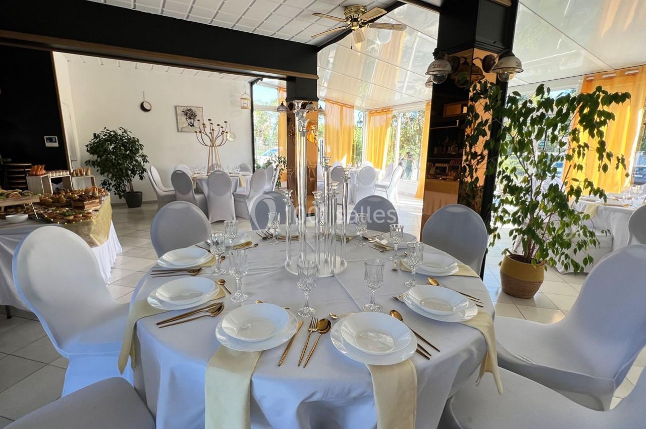 Salle de réception lumineuse avec tables rondes dressées, nappes blanches, couverts dorés et décorations élégantes.