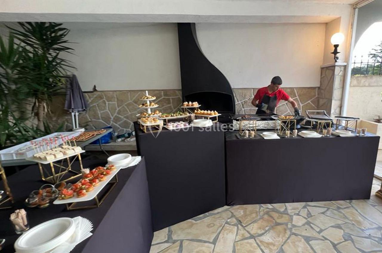 Un chef prépare des plats derrière un buffet garni de verrines, amuse-bouches et assiettes dans un espace extérieur couvert.