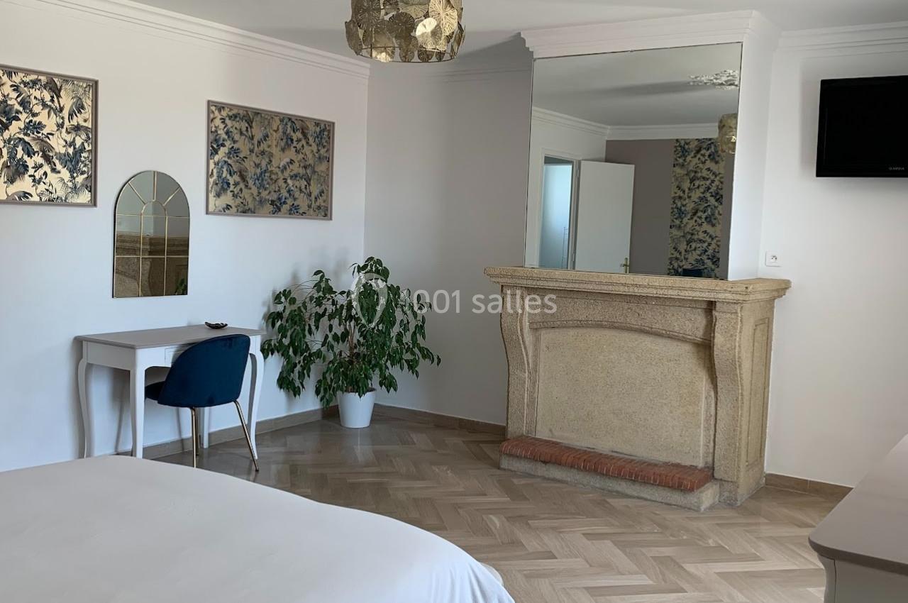 Chambre lumineuse avec parquet, bureau, chaise bleue, cheminée en pierre, miroir et décoration murale florale.