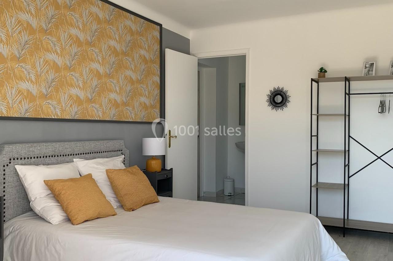 Chambre lumineuse avec lit double, coussins jaunes, tableau à motifs végétaux et étagère en métal noir.