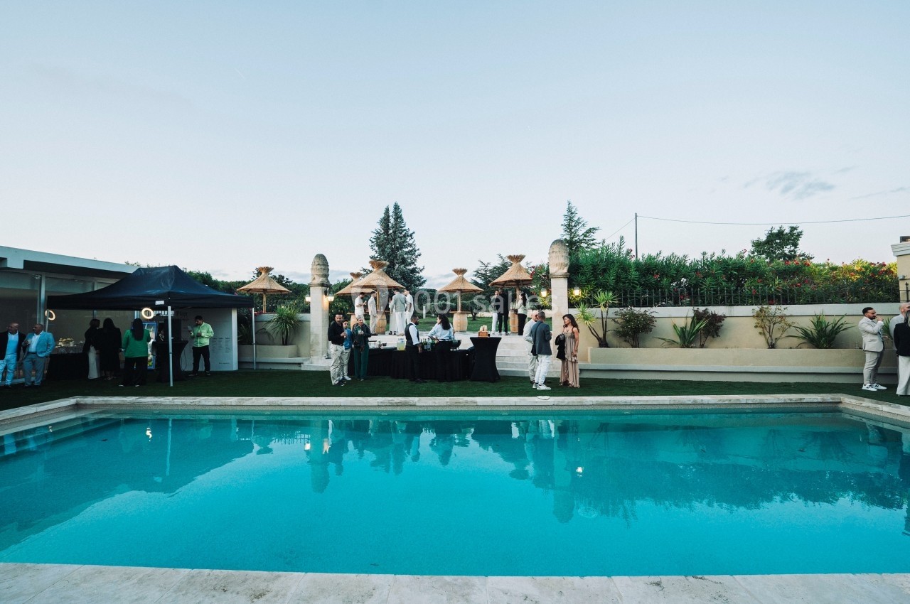 Personnes rassemblées près d'une piscine dans un jardin, avec des tables et des décorations en arrière-plan.