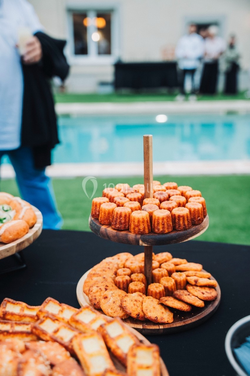 Plateau de cannelés et biscuits apéritifs disposés sur une table près d'une piscine en extérieur.