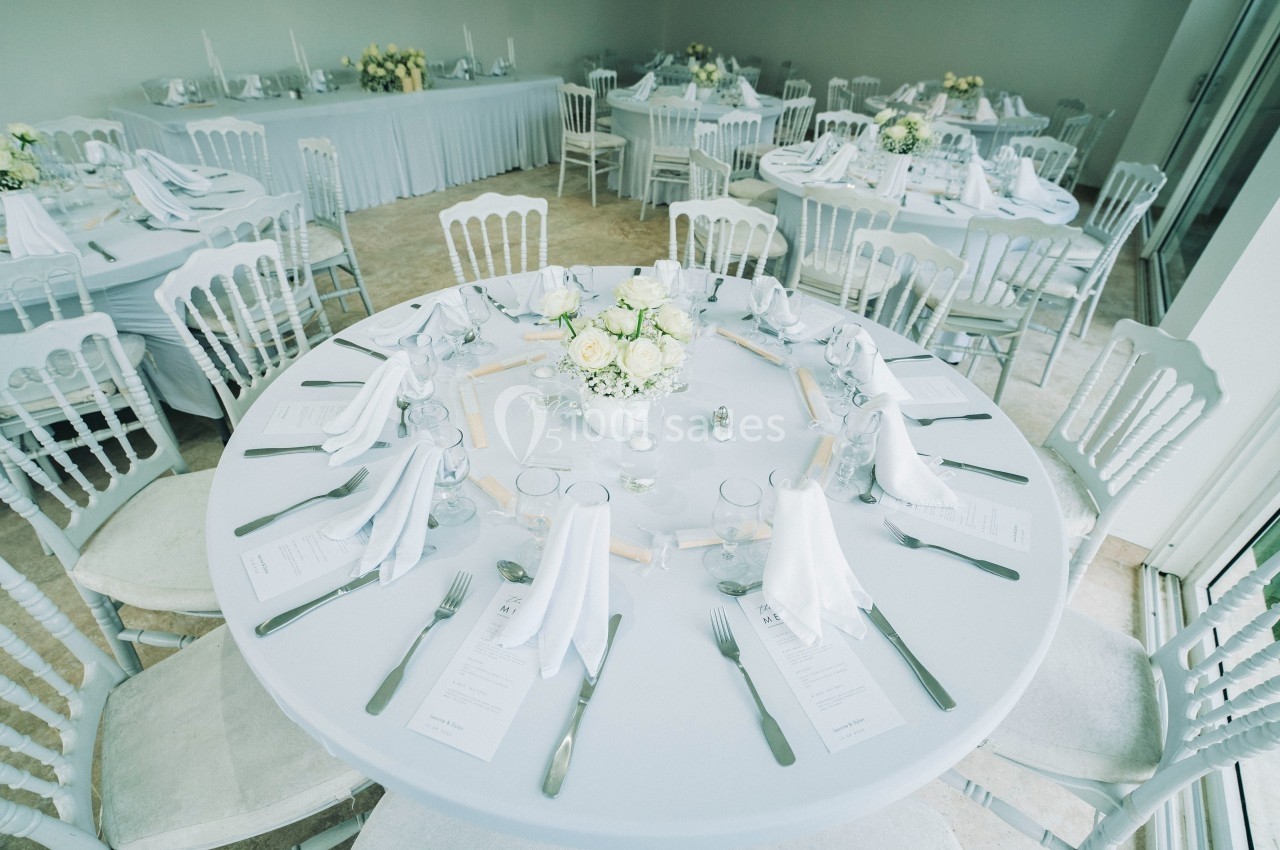 Salle de réception avec tables rondes dressées, nappes blanches, serviettes pliées et centre de table floral.