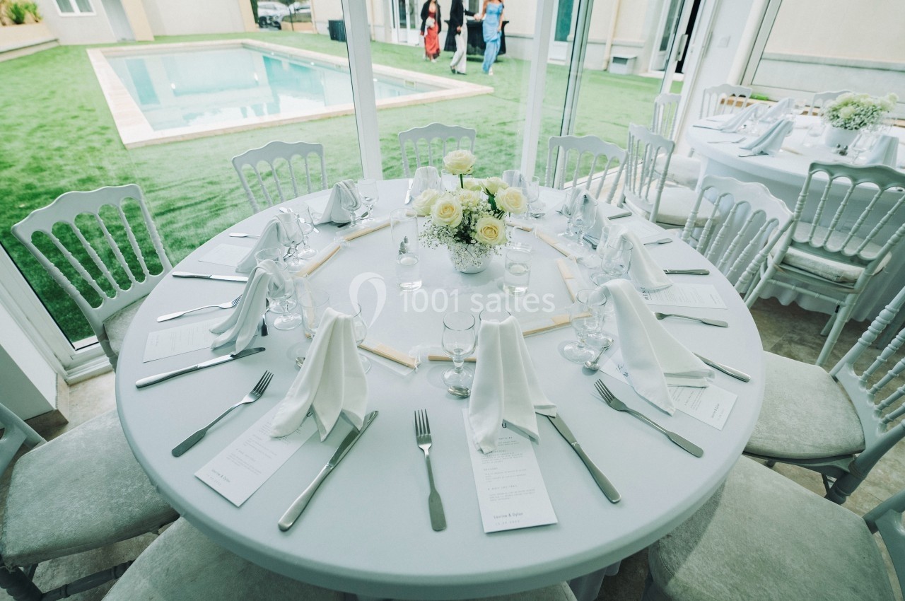 Table ronde dressée avec nappes blanches, couverts, serviettes pliées et un centre de table floral, près d'une piscine.