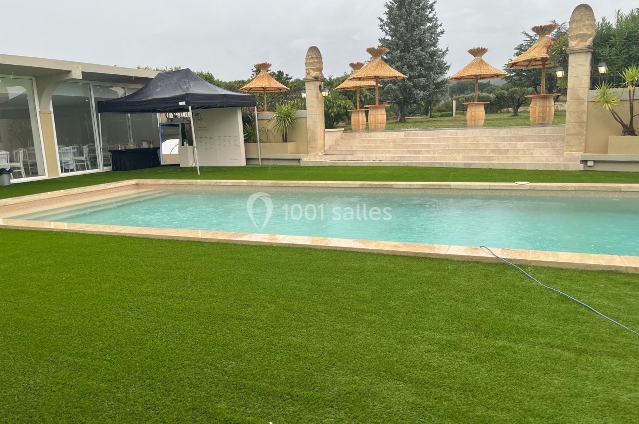 Piscine extérieure entourée de gazon artificiel, avec des parasols en paille et des colonnes en arrière-plan.