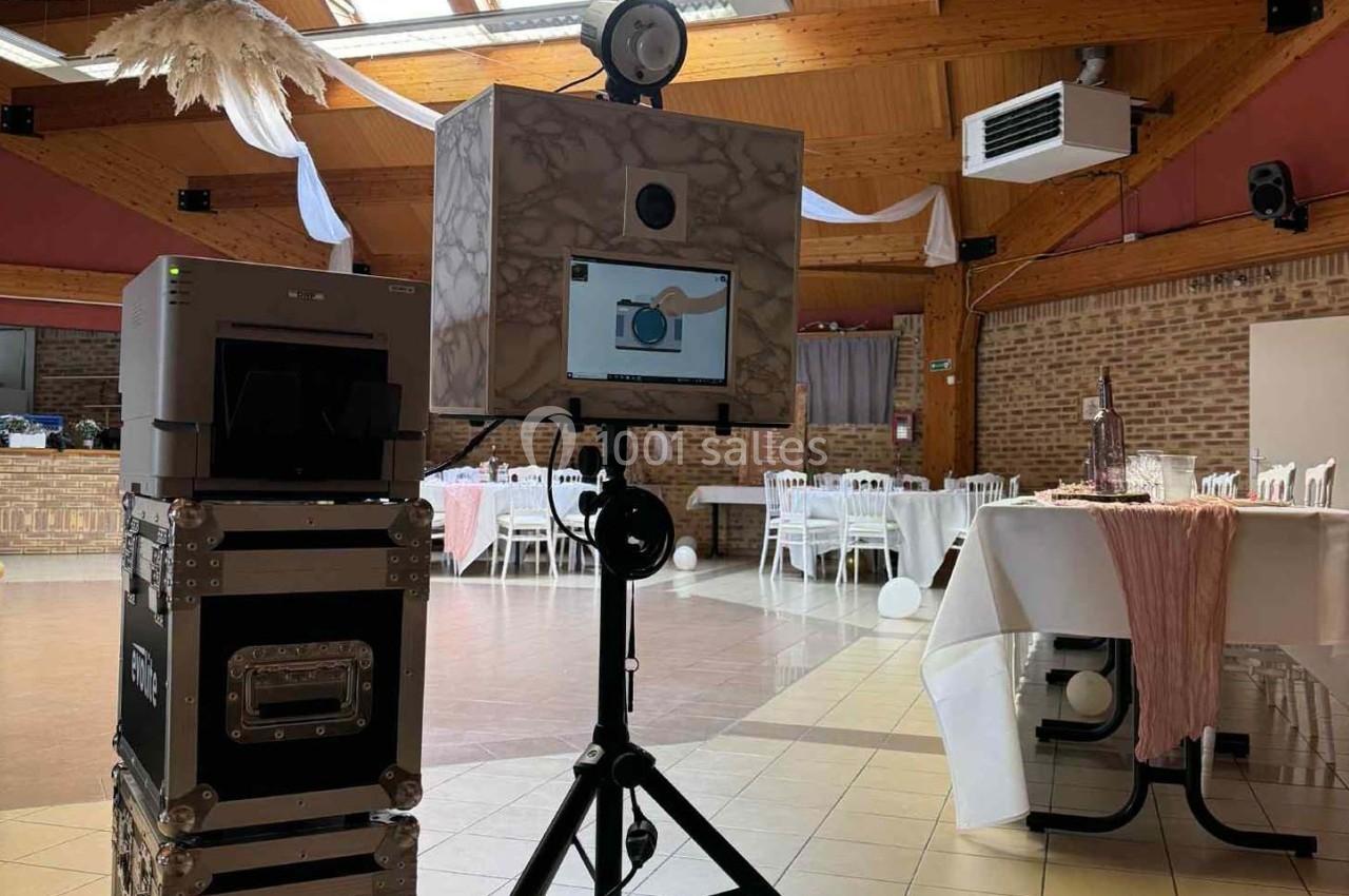 Borne photo installée dans une salle de réception avec tables décorées et lumière naturelle entrant par le toit.