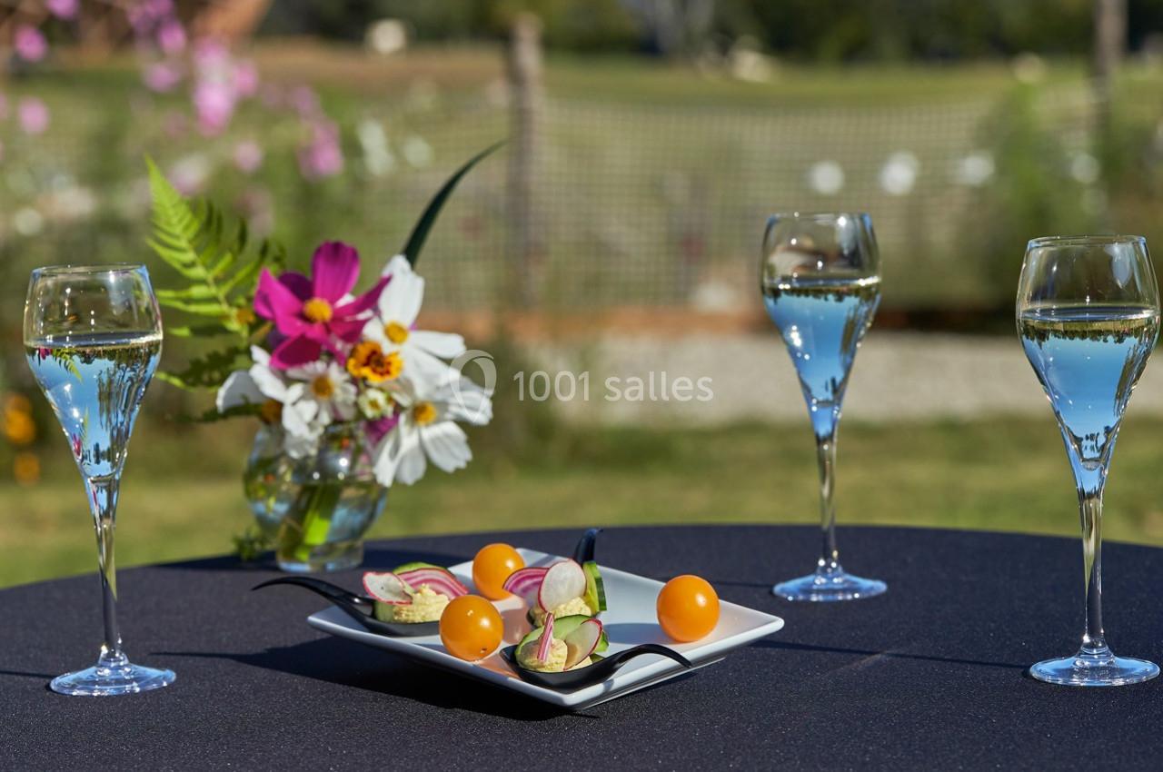 Trois verres de vin pétillant entourent une assiette de légumes et fleurs comestibles sur une table en extérieur.