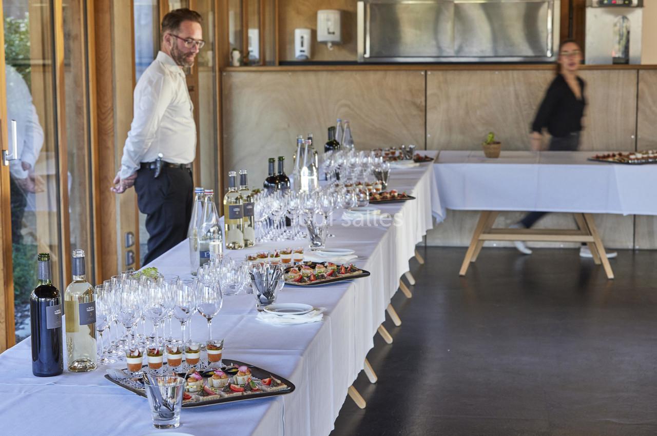 Table dressée avec verres, bouteilles et plateaux de canapés dans une salle lumineuse, un homme et une femme en arrière-plan.