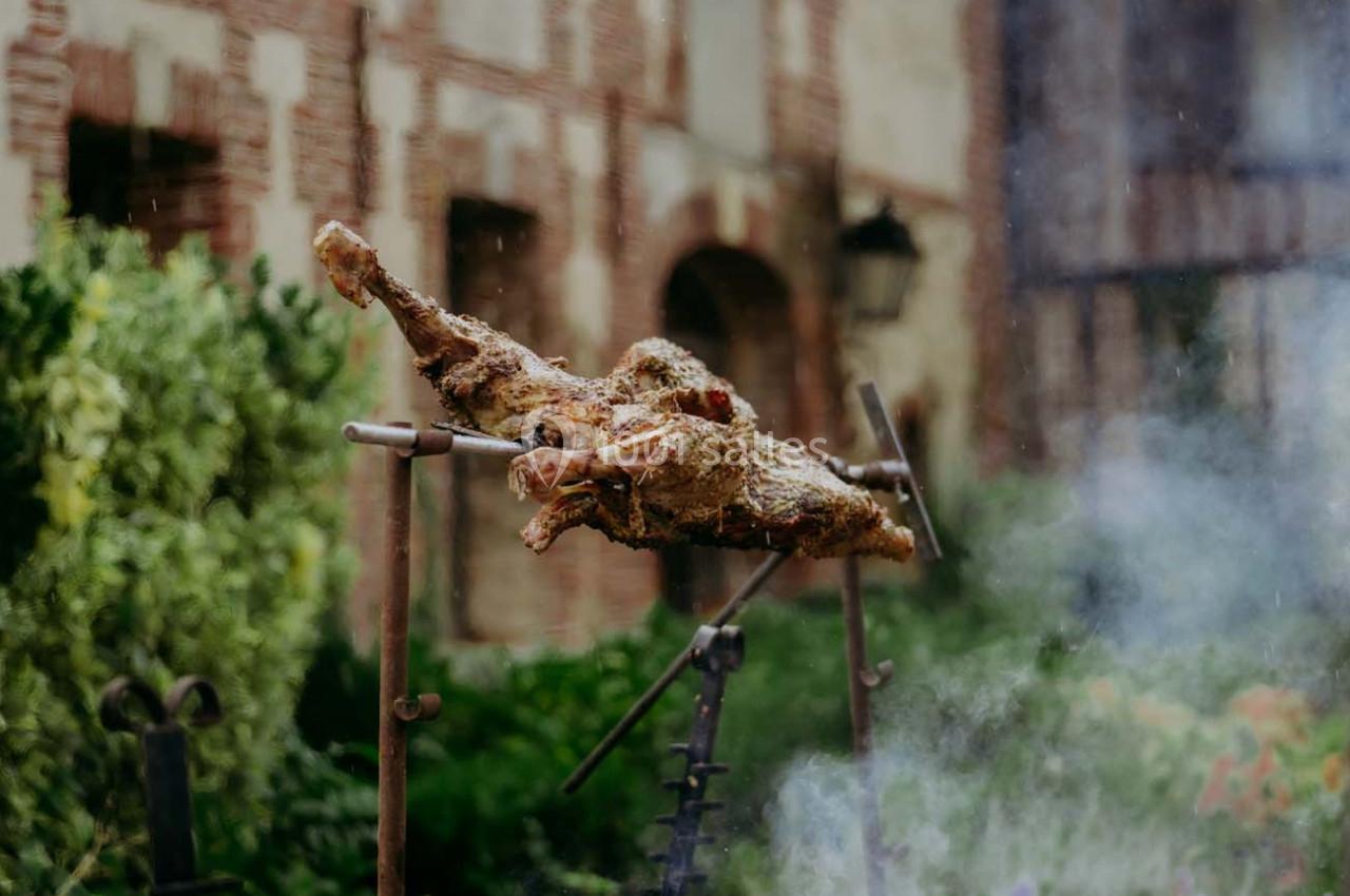 Viande rôtie sur une broche en extérieur, devant un mur en briques et un jardin verdoyant.