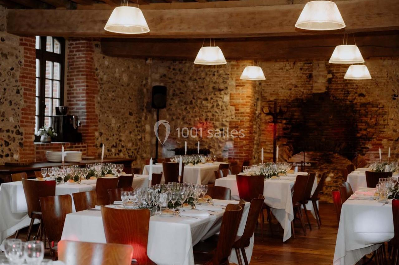 Salle de réception avec murs en pierre, tables dressées avec nappes blanches, chaises en bois et éclairage suspendu.
