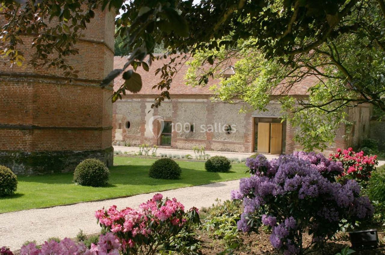 Jardin fleuri avec allée gravillonnée, buissons taillés, rhododendrons en fleurs et bâtiments en briques en arrière-plan.