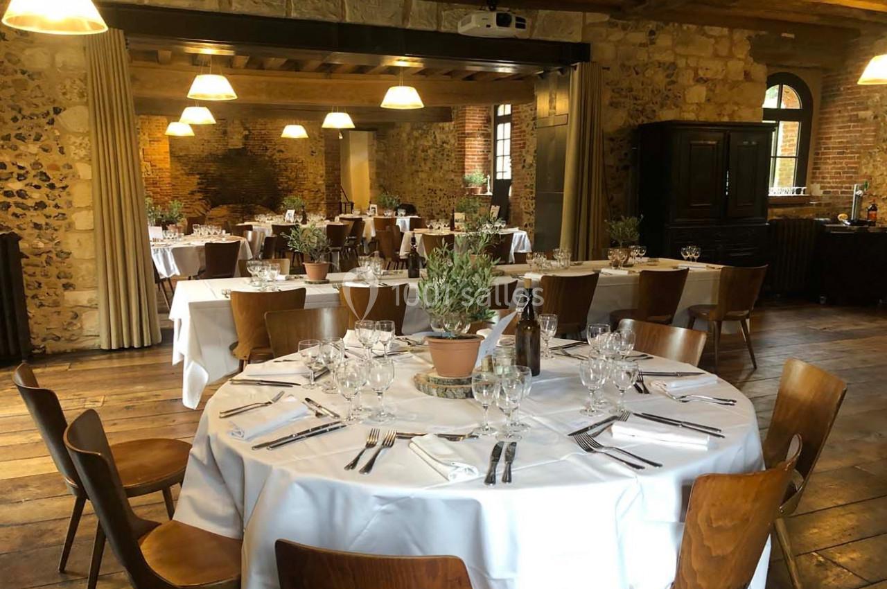 Salle de réception avec tables rondes dressées, nappes blanches, chaises en bois et décor rustique en pierre et bois.