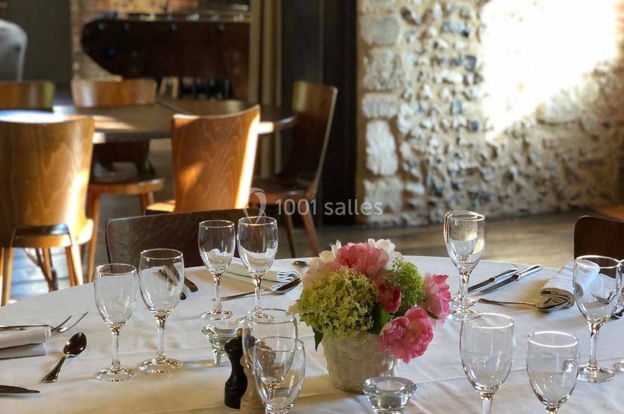 Table dressée avec nappes blanches, verres, couverts et bouquet de fleurs, dans une salle lumineuse aux murs en pierre.