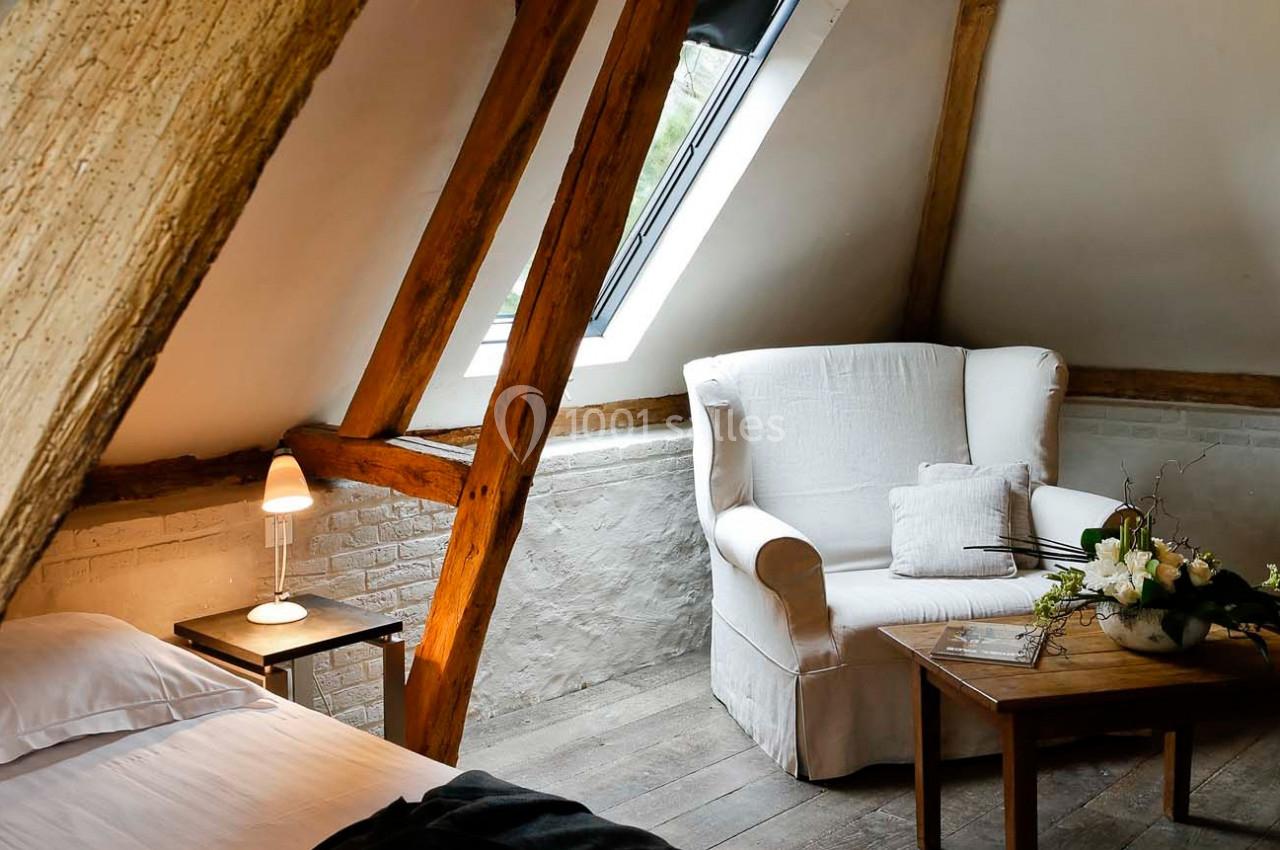 Fauteuil blanc près d'une table en bois, sous des poutres apparentes, avec une lampe et un lit à proximité.