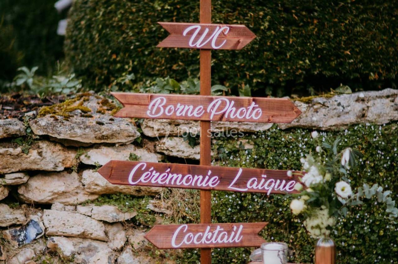 Panneau en bois avec flèches indiquant WC, borne photo, cérémonie laïque et cocktail, devant un mur en pierre.