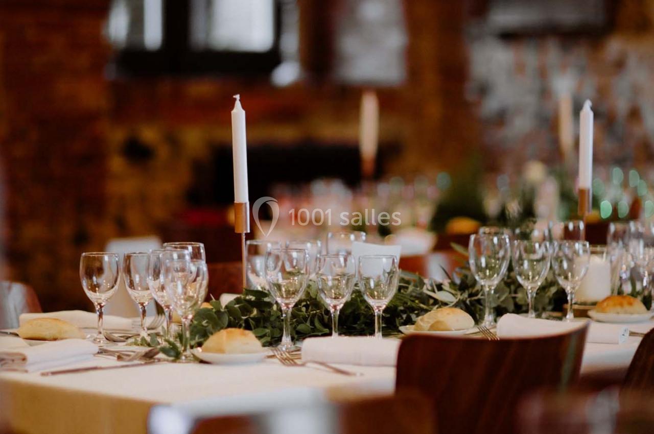 Table dressée avec nappes blanches, verres, bougies et décorations végétales dans une salle au mur de briques.