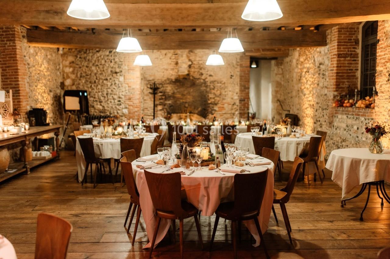 Salle de réception chaleureuse avec murs en pierre, tables rondes dressées et éclairage suspendu.