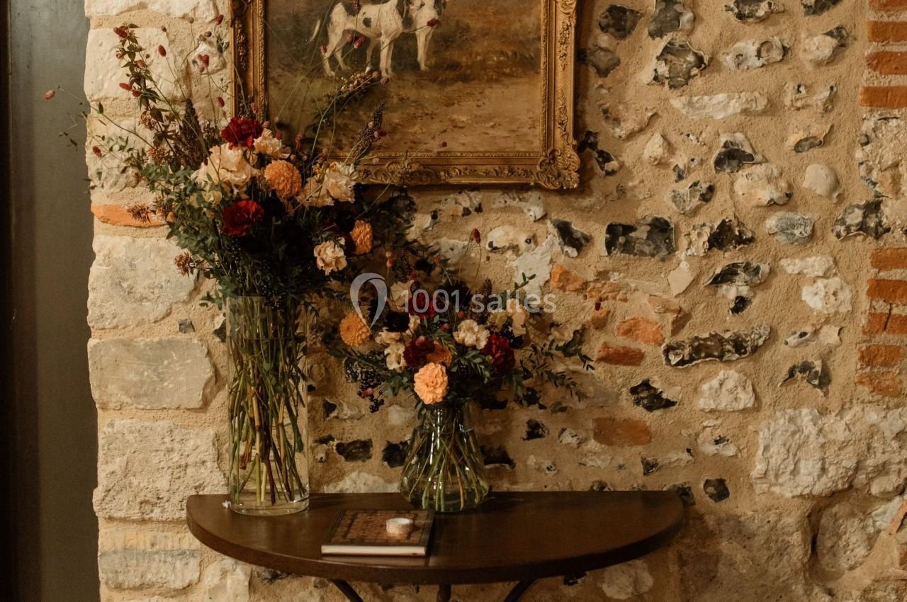 Table en bois avec deux bouquets de fleurs devant un mur en pierre, surmontée d'un tableau représentant des chiens.
