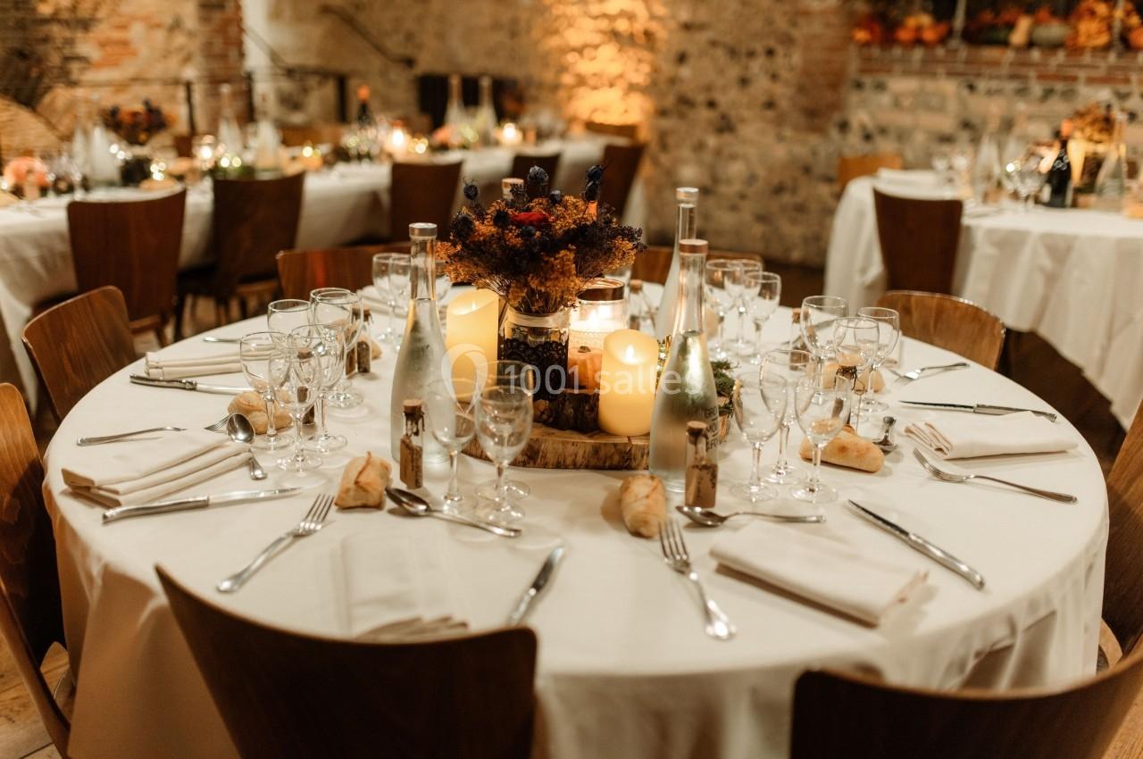 Table ronde élégamment dressée avec nappes blanches, couverts, bougies et décoration florale dans une salle en pierre.