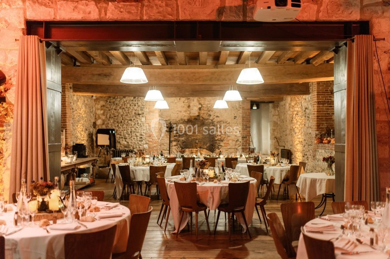 Salle de réception chaleureuse avec tables rondes dressées, éclairage doux et murs en pierre apparente.