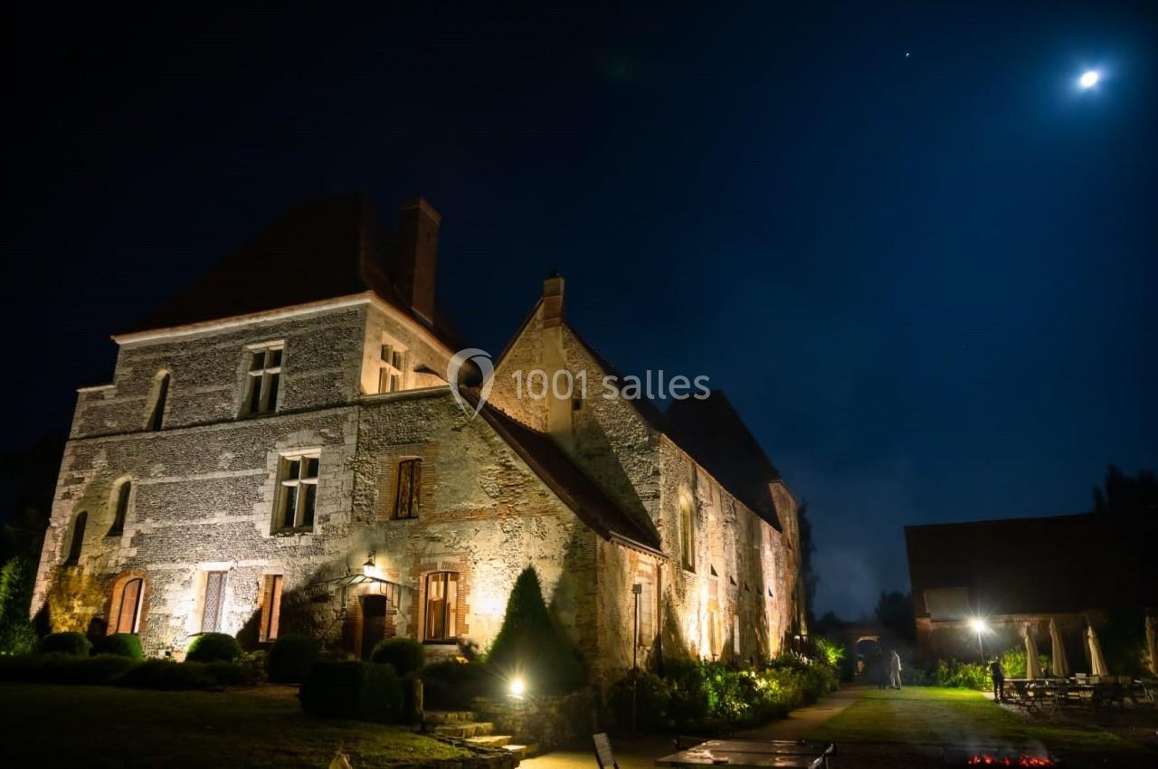 Manoir en pierre éclairé la nuit, entouré de végétation, sous un ciel étoilé avec une lune brillante.