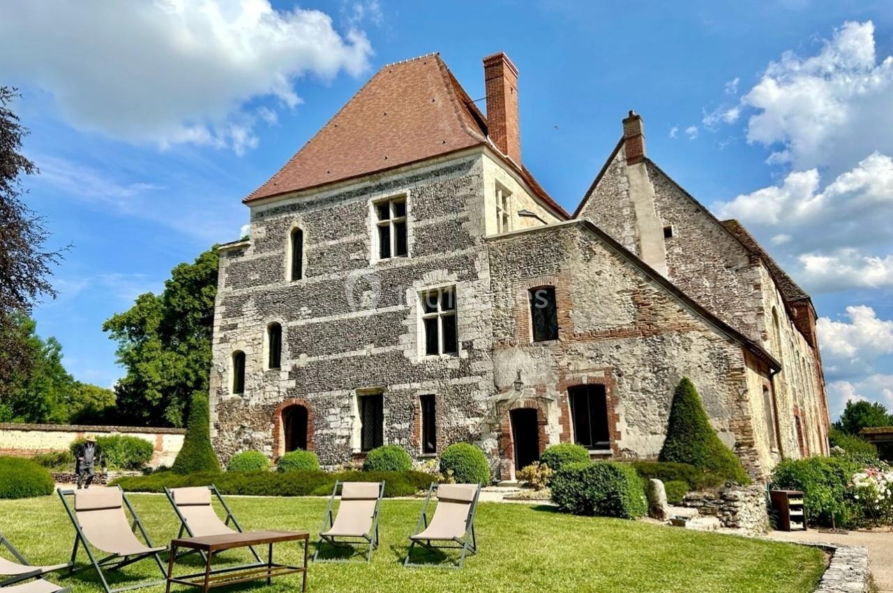 Manoir en pierre avec toit en pente, entouré d'un jardin aménagé et de chaises longues sous un ciel partiellement nuageux.