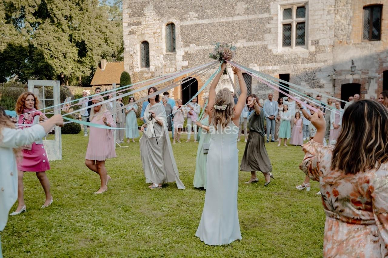 Des personnes en tenue élégante dansent en cercle à l'extérieur, tenant des rubans colorés attachés à un bouquet.