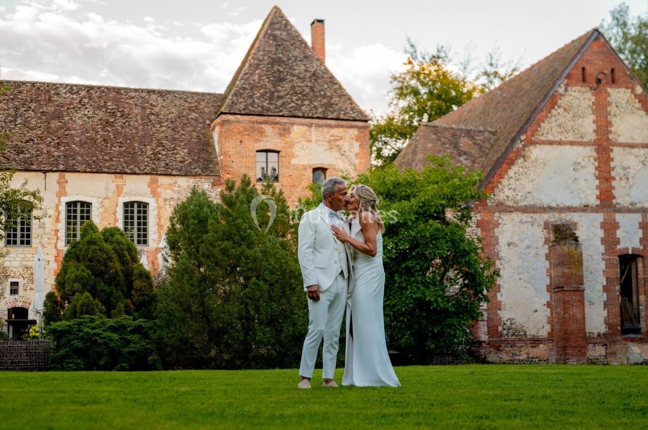 Un couple en tenue élégante se tient tendrement dans un jardin devant un bâtiment ancien en pierre.