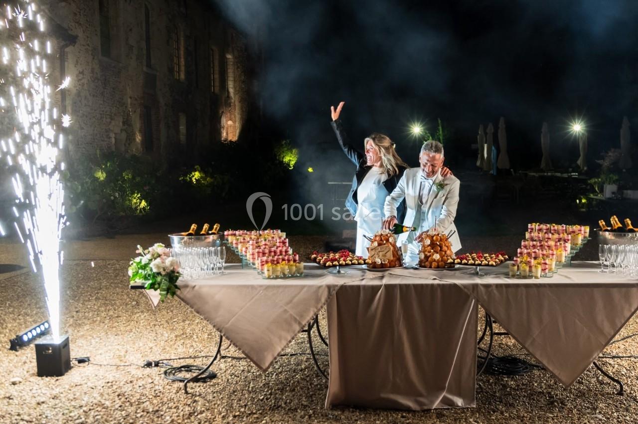 Un couple célèbre un événement devant une table garnie de desserts et de champagne, avec des feux d'artifice en arrière-plan.