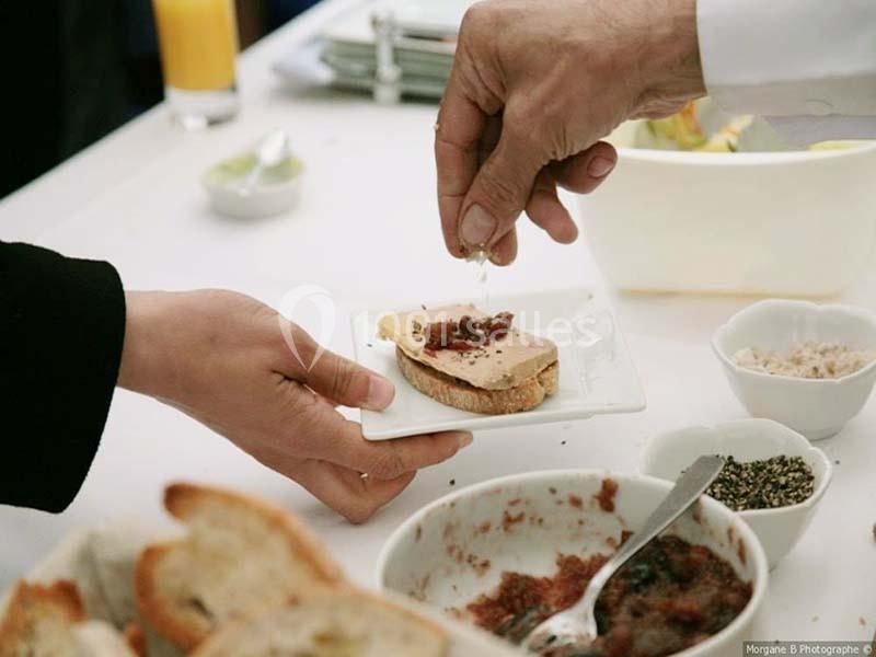 Une personne ajoute une garniture sur une tranche de pain avec du foie gras, entourée de condiments sur une table.