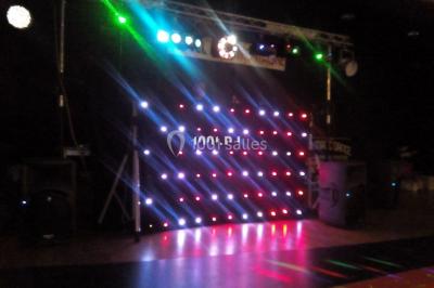 Un DJ mixe derrière une table éclairée de lumières LED violettes, avec un mur décoratif en arrière-plan.