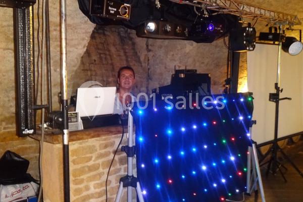 Un DJ installé derrière une table équipée de lumières LED colorées et d'équipements audio dans une salle en pierre.