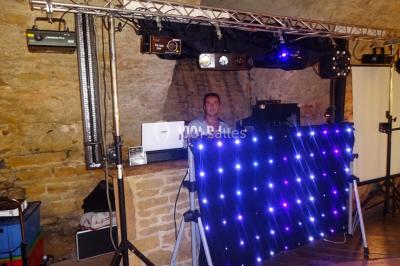 Un homme souriant se tient près d'une table de mixage éclairée, avec un écran projetant une vidéo en arrière-plan.