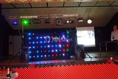 Un homme souriant se tient près d'une table de mixage éclairée, avec un écran projetant une vidéo en arrière-plan.