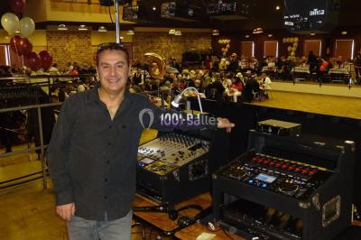 Un homme souriant se tient près d'une table de mixage éclairée, avec un écran projetant une vidéo en arrière-plan.