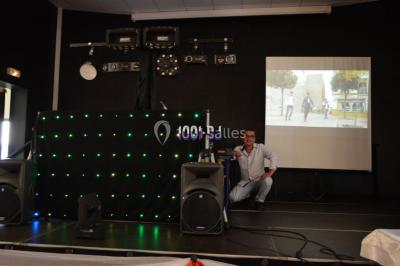 Un homme souriant se tient près d'une table de mixage éclairée, avec un écran projetant une vidéo en arrière-plan.